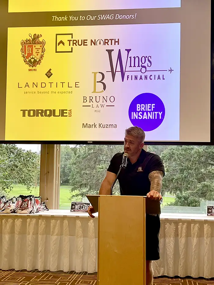 Stephen Foertsch at the podium at the Minnesota 100 Club golf tournament dinner, under a slide thanking donors including Bruno Law